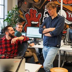 Drie collega's in gesprek bij hun bureaus met beeldschermen voor een kleurrijke graffitimuur