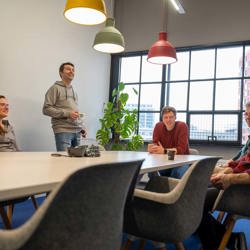 Vijf collega's in een lachend gesprek rond een vergadertafel onder gekleurde hanglampen