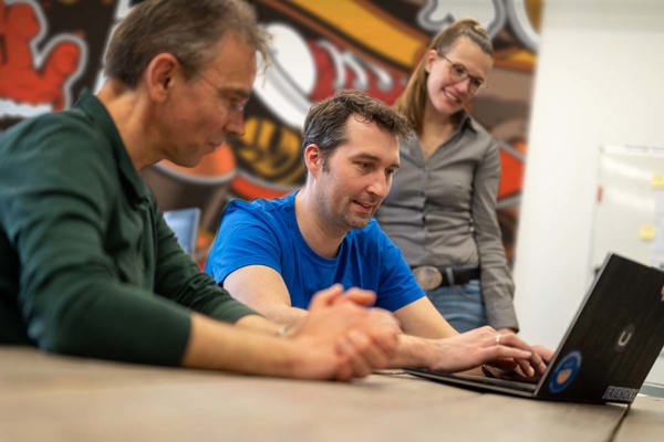 Drie collega's kijken samen naar een laptop aan tafel voor een kleurrijke graffitimuur